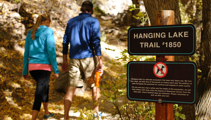 Hanging Lake Information Immersed in the serene ambiance of the Hanging Lake Trail, two hikers move past the Hanging Lake sign, ascending the dirt trail.