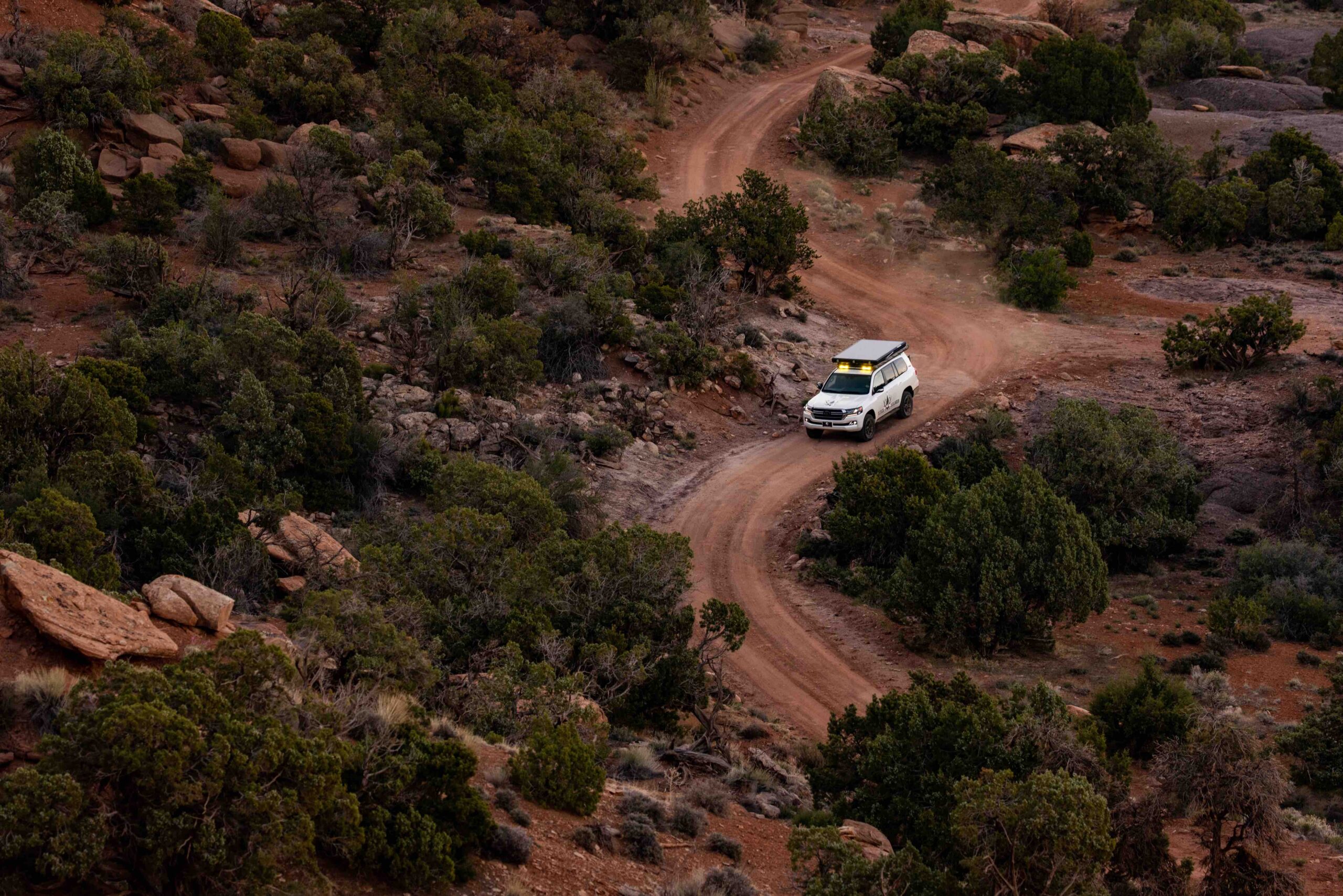 Colorado Overlander Info Photo Captured from above, this aerial photo showcases the Colorado Overlander's Land Cruiser confidently navigating a vibrant red dirt road. Surrounded by piñon trees, the dust clouds billow up from behind the tires, accentuating the adventurous spirit of the journey. The dynamic scene captures the essence of off-road exploration and the rugged beauty of the terrain beneath the Land Cruiser's wheels.