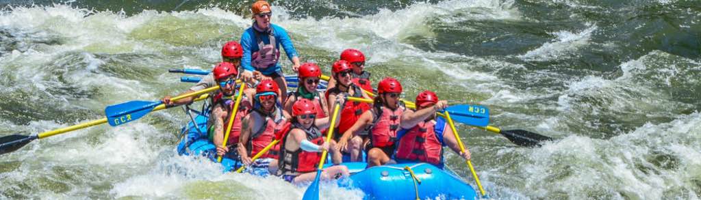 GCR Rafting–1400×400 In this captivating image, a guide expertly navigates a full boat of guests through the exhilarating Shoshone Rapids. Adorned in essential safety gear, including life jackets and helmets, the group paddles together, showcasing teamwork and skill as they conquer the rapids.