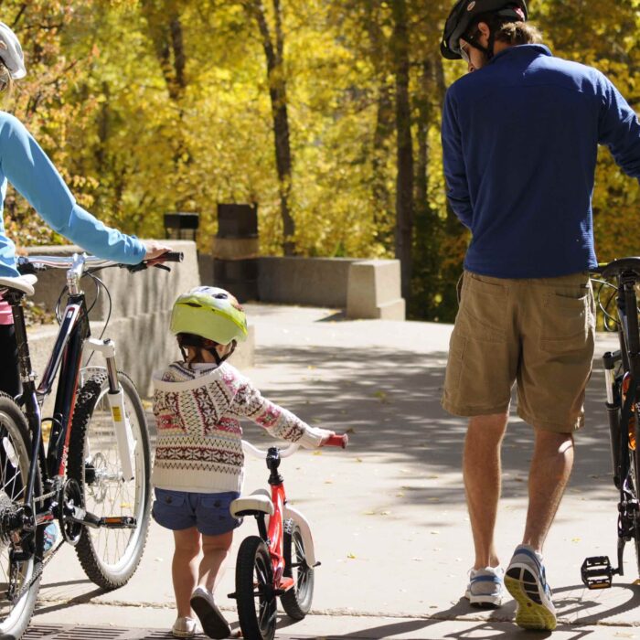 In this charming image, two adults and a child walk their bikes along a paved biking trail, donned in helmets, athletic clothing, and comfortable tennis shoes.
