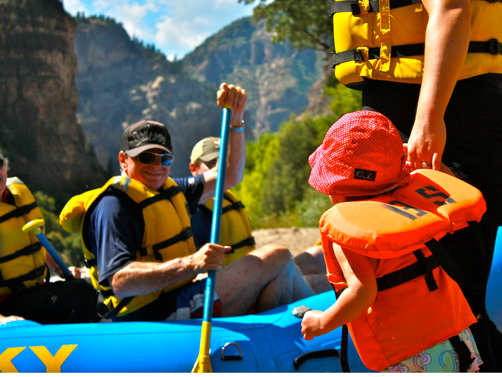 In this photo, a young child and her mother hold hands and greet a boat of rafters at Grizzly Creek beach. They all wear bright colored safety gear and hats.