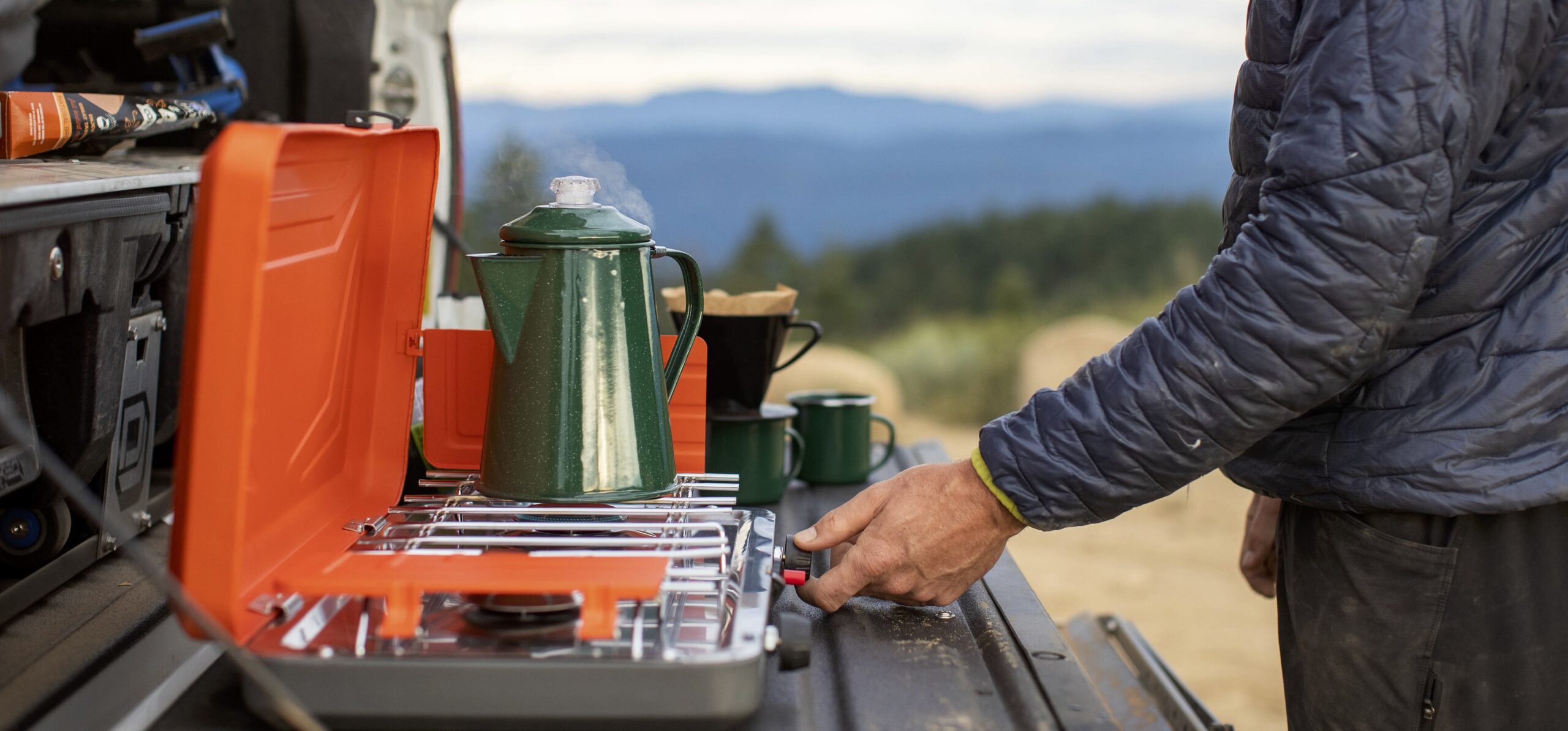 In this image, a hand turns on a camp stove to heat water for pour-over coffee, setting the stage for a tranquil morning camping experience in the great outdoors. The up-close shot focuses on the action, withholding the identity of the person behind the scene.