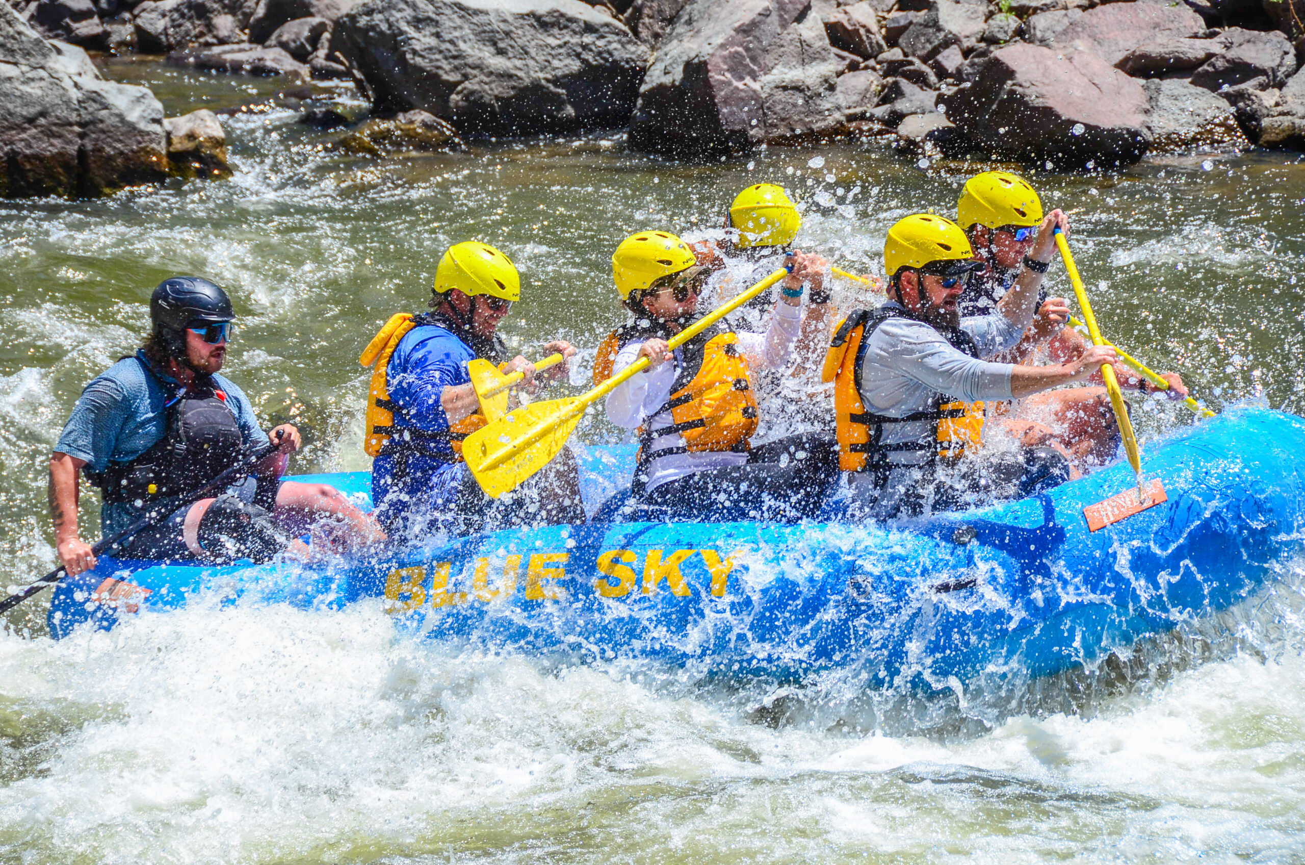In this compelling image, a proficient guide expertly directs a fully occupied boat from the rear as participants paddle through the whitewater together. Cloaked in life jackets and helmets, the group showcases seamless teamwork, with water splashing around them, capturing the dynamic and exhilarating essence of their adventure.