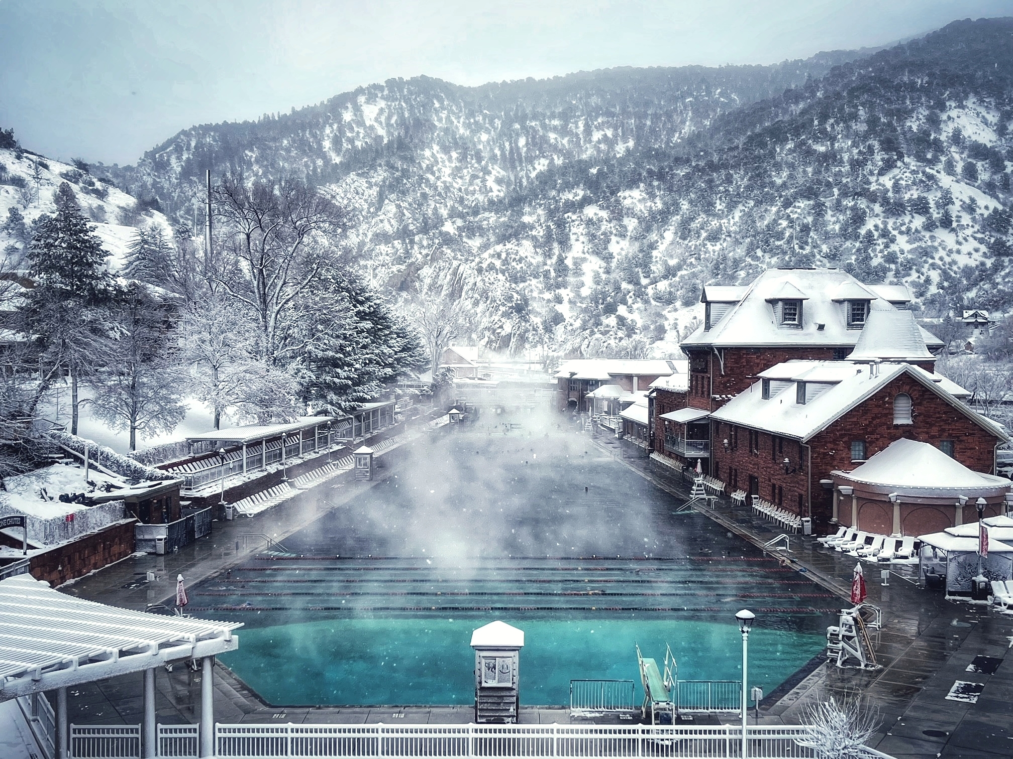 Scenic winter view of Glenwood Hot Springs pool, surrounded by snow-covered mountains and buildings. The soothing hot springs mineral waters create a tranquil scene with steam rising, offering a serene contrast to the snowy landscape.