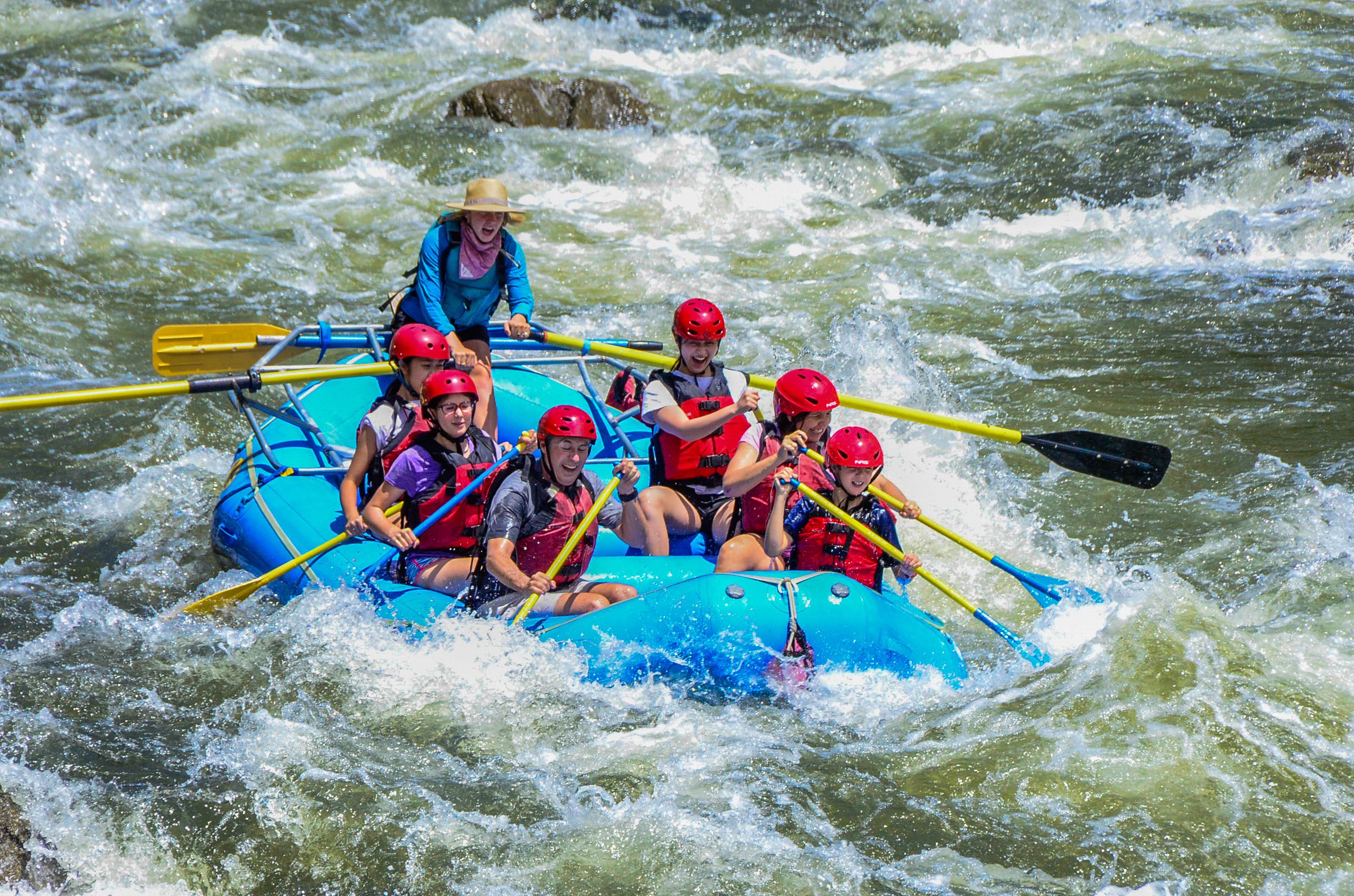 This is an action photo of a group of people and their guide paddling through rapids on the Colorado River. They wear safety gear as they get splashed, wearing big smiles on their faces.